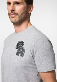 Szara bawełniana koszulka z krótkim rękawem, z czarnym graficznym logo "PR" na lewej piersi, gładka faktura, okrągły dekolt.