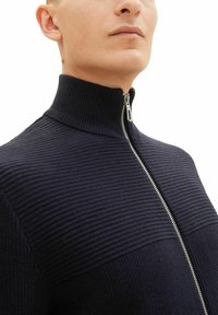 Sweater zippé marine à côtes avec col montant ; présente des rayures verticales, une fermeture éclair métallique et une texture douce, conçu pour une apparence ajustée.