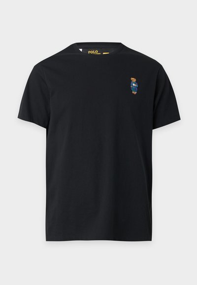 Μαύρο T-shirt με κοντά μανίκια και στρογγυλή λαιμόκοψη με μικρό κεντητό αρκουδάκι που φοράει μπλε σαλόπετα στα αριστερά του στήθους.