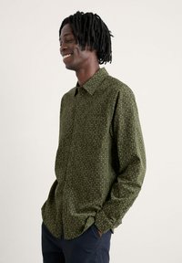 Camicia verde con bottoni, con una superficie testurizzata, caratterizzata da un piccolo motivo a pois bianchi e blu, e una tasca sul petto.