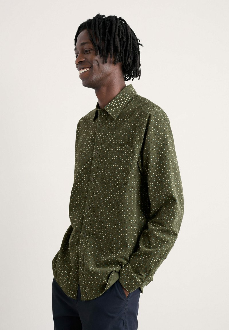 Camicia verde con bottoni, con una superficie testurizzata, caratterizzata da un piccolo motivo a pois bianchi e blu, e una tasca sul petto.