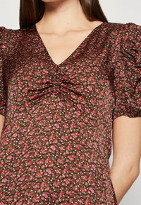 Blouse fleurie en tissu foncé avec des motifs de fleurs roses et rouges. Elle présente un décolleté en V et un détail froncé à la poitrine, avec des manches bouffantes.