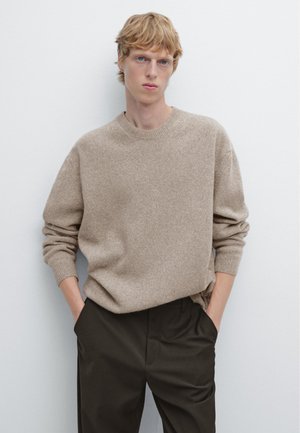 Beige, gemarmerde brei sweater met een ronde hals en verlaagde schouders, gedragen over donkere broek, met handen in de zakken voor een effen achtergrond.