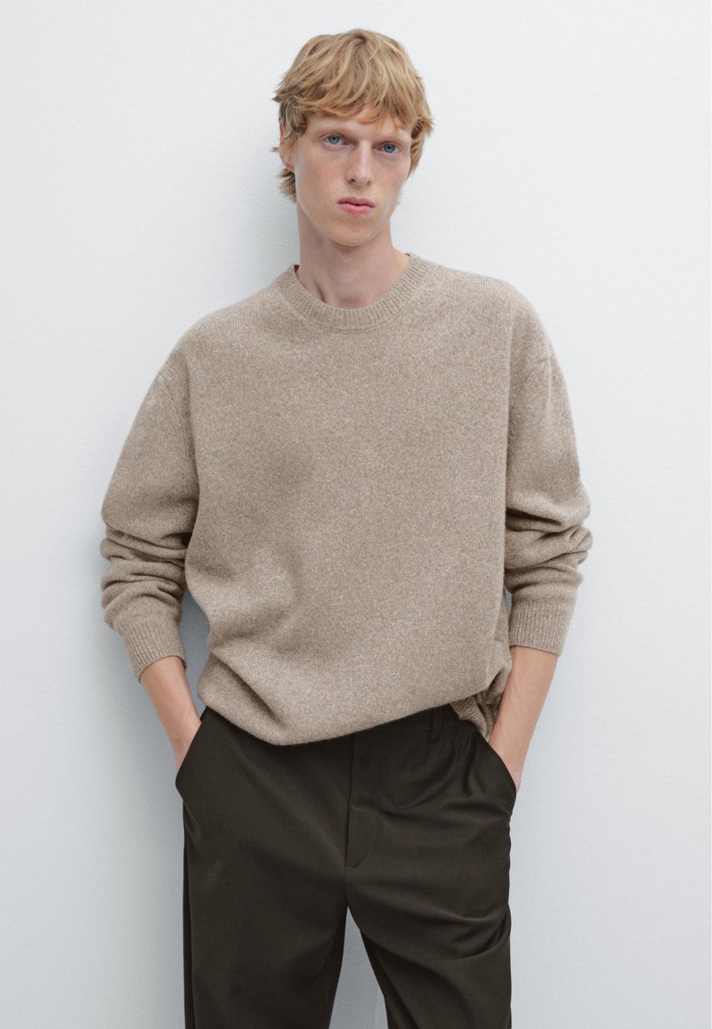 Beige, texturerad stickad tröja med rund halsringning och sänkt axel, bärs över mörka byxor, med händerna i fickorna mot en enkel bakgrund.