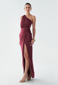 Bordové jedno ramenové šaty z jemnej látky, s drapériou a rozparkom nad stehno. Kombinované so zlatými sandálmi na remienky.