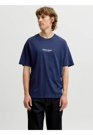 Młody mężczyzna o jasnobrązowych włosach, ubrany w granatowy T-shirt Jack & Jones i czarne spodnie, stoi na tle jednolitego jasnego tła.