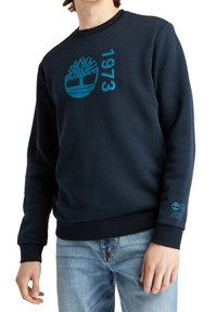 Sudadera azul marino de algodón con cuello acanalado, que presenta un logo de árbol azul y "1973" impreso en el pecho. Tejido texturizado.