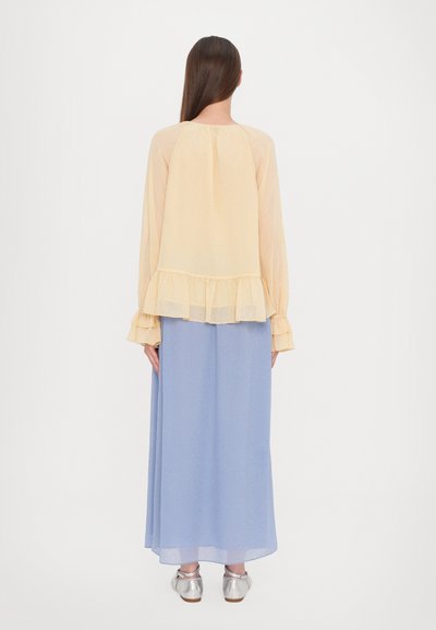 Geel chiffon blouse met een gerimpelde onderrand en mouwen, gecombineerd met een lange lichtblauwe rok. De outfit heeft een lichte textuur.