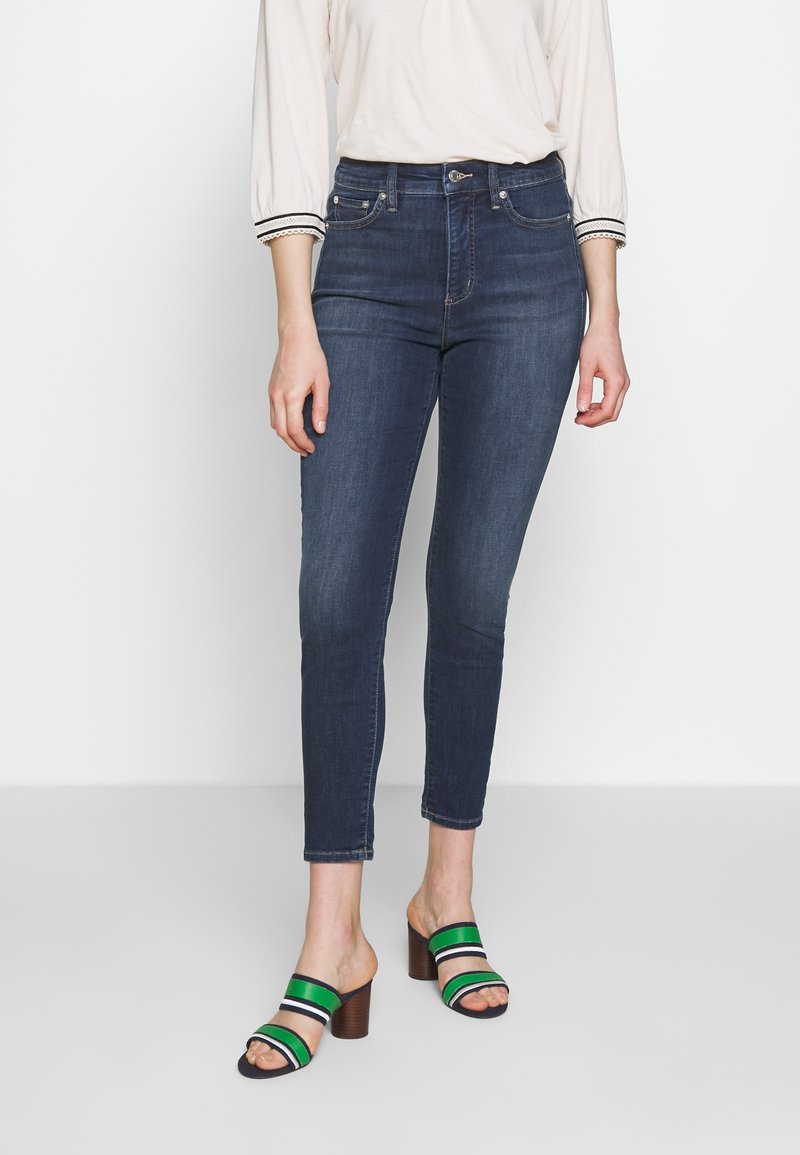 Dark blue skinny jeans with a high waist and a smooth texture, paired with black and green striped heeled sandals.