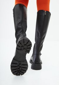 Persona que lleva botas negras hasta la rodilla con suelas gruesas y leggings naranjas, mostrando el patrón de la suela sobre un fondo blanco.