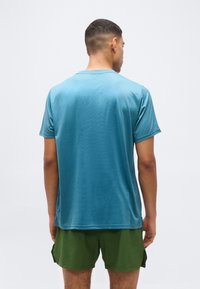 Άνδρας που φοράει μπλουζάκι με  κοντά μανίκια τιρκουάζ και πράσινα σορτς, στέκεται με την πλάτη προς τα εμπρός σε απλό λευκό φόντο.