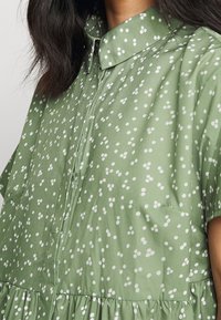 Femme portant une robe verte claire à manches courtes avec un motif de points blancs groupés et un col pointu, apparente des épaules à la taille.