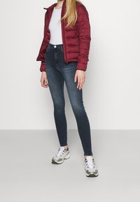 Czerwona kurtka puchowa, dopasowana biała koszulka, ciemne jeansy skinny i szare buty sportowe z żółtymi akcentami, stojąca na jasnoszarym tle.