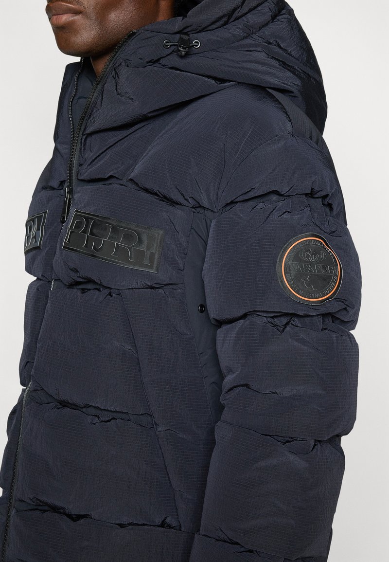 Veste rembourrée bleu marine avec fermeture éclair, grandes poches avant, texture matelassée et écussons logo sur la poitrine et la manche.