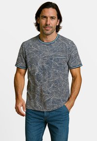 Man met schouderlang donker haar draagt een blauw-wit t-shirt met bladerenpatroon en een blauwe spijkerbroek, staat met één hand in de zak.