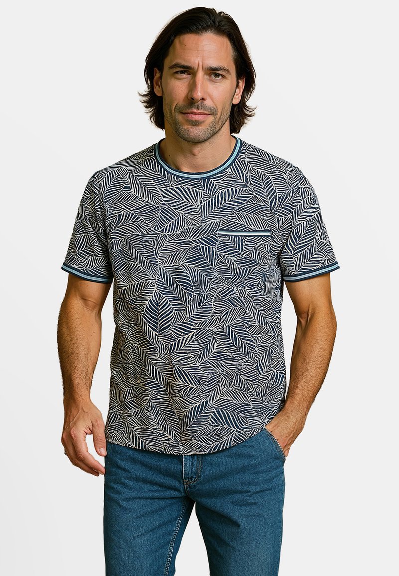 Man met schouderlang donker haar draagt een blauw-wit t-shirt met bladerenpatroon en een blauwe spijkerbroek, staat met één hand in de zak.