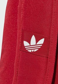 La tela roja presenta un parche de logo blanco con tres hojas y tres franjas horizontales. La textura parece suave con bordes reforzados.