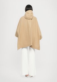 Poncho beige avec capuche, présentant un design ample et une texture douce, associé à un pantalon plissé blanc et des talons beiges.