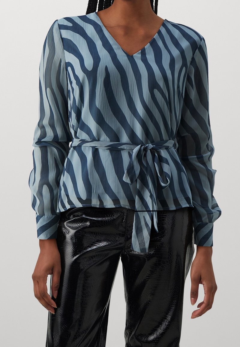 Femme portant un chemisier à manches longues rayé zèbre bleu et marine avec ceinture à la taille et un pantalon noir brillant et texturé.