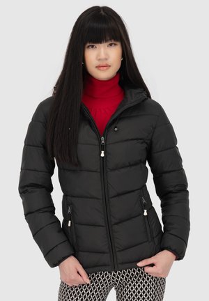 Jeune femme aux longs cheveux noirs portant une doudoune noire sur un col roulé rouge et un pantalon à motifs, se tenant devant un fond uni.