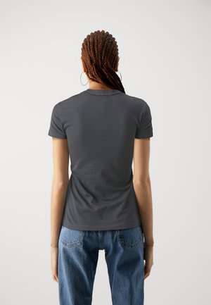 Kvinde med flettet hår iført creol-øreringe, en mørkegrå T-shirt med korte ærmer og blå jeans, står vendt mod en ensfarvet lys baggrund.
