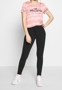 Pink tie-dye t-shirt with black "Hollister" logo, paired with black leggings and white sneakers. Sleek, fitted design with elastic waistband.