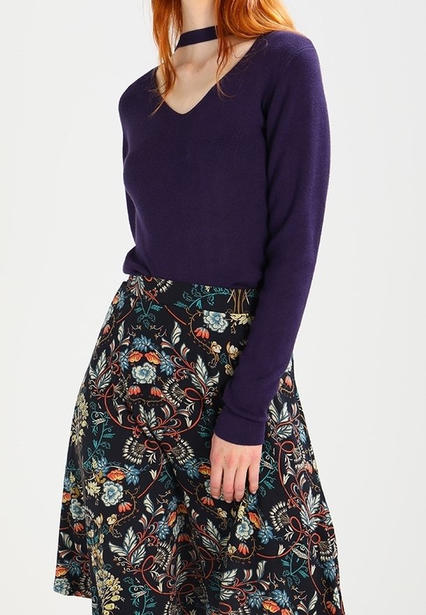 Femme portant un pull violet à manches longues et col en V et une jupe midi noire à motif floral avec des fleurs rouges, bleues et beiges.