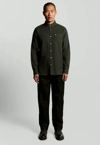 Chemise boutonnée vert foncé avec col, poche poitrine simple et petit logo. Associée à un pantalon noir et des chaussures noires texturées.