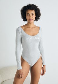 Šedý bodysuit s dlouhým rukávem a výstřihem ve tvaru lodi, má přiléhavý střih; na hrudi se nachází logo "GUESS" v bílé barvě. Hladká textura látky.