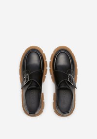 Chaussures en cuir noir avec une semelle en caoutchouc épaisse et brune, dotées d'une sangle à boucle et d'un bout arrondi. Texture lisse et design minimaliste.