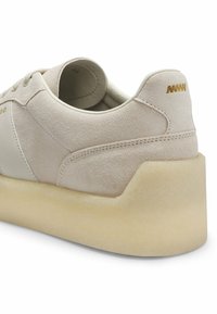 Baskets en daim beige avec semelle épaisse en crêpe blanc cassé, coutures minimales et petit logo doré sur le talon, vues sous un angle arrière latéral.