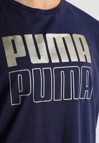 Námořnická modrá bavlněná tričko s velkým stříbrným textem "PUMA" ve dvou řadách. Krátké rukávy a kulatý výstřih s jemnou strukturou.