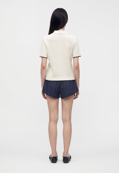 Femme aux longs cheveux foncés portant un haut en maille blanc à manches courtes et un short bleu marine, vue de dos, debout sur un fond uni.