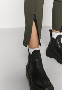 Olivgröna tajta leggings med sidodragkedjor, kombinerat med svarta läderankelstövlar med tjock sula och elastiska insatser.