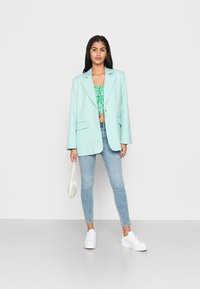 Blazer oversized verde menta com lapelas em bico, dois bolsos frontais, usado sobre uma blusa curta verde estampada, combinado com jeans azul claros e ténis brancos.