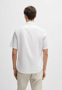 Camicia bianca a maniche corte, tessuto di cotone, colletto classico, orlo curvato, design semplice senza motivi o accenti visibili. Vista posteriore mostrata.
