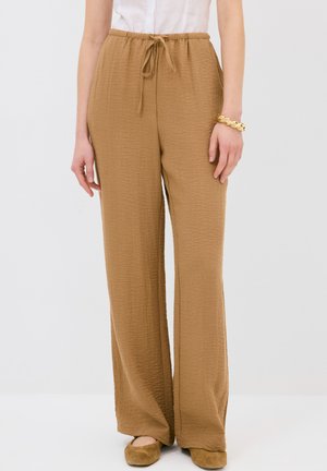Pantaloni a gamba larga in un tessuto marrone chiaro e strutturato. Presentano una vita elasticizzata con cordoncino. Completati da scarpe marroni morbide.