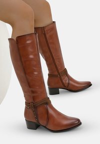Botas marrones hasta la rodilla, fabricadas en cuero liso, con cremallera lateral, una cinta texturizada con acentos metálicos y un tacón bajo. 