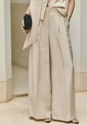 Pantalones beige de tiro alto y pierna ancha combinados con abrigo sin mangas a juego, clutch negro tejido, pulsera dorada y sandalias negras abiertas en los dedos.