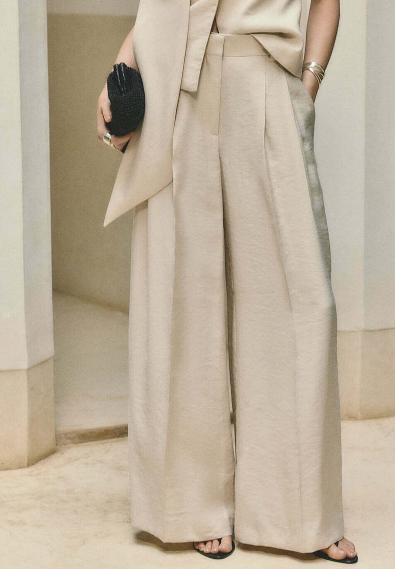 Pantalon beige taille haute à jambes larges associé à un manteau sans manches assorti, pochette noire tissée, bracelet doré et sandales noires à bouts ouverts.