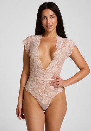 Vrouw draagt een lichtroze kanten body met korte mouwen en een diepe V-hals, staat met één hand op haar heup tegen een effen achtergrond.