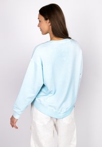 Light blue sweatshirt with a relaxed fit, ribbed cuffs, and waistband. Features subtle heart detail printed on the back. Soft fabric texture.