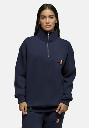Femme portant un pull bleu marine avec un col zippé haut et un pantalon assorti, avec de petits détails de boutons orange et or.