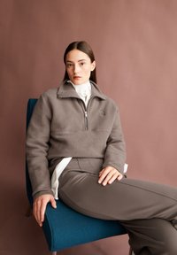 Grijze fleece pullover met een hoge kraag en halve rits, gecombineerd met grijze op maat gemaakte broeken. Model zit op een blauwe stoel tegen een beige achtergrond.