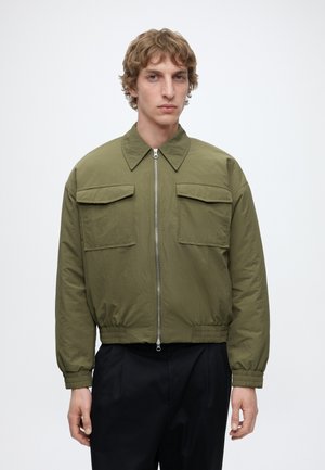 Jeune homme portant une veste zippée vert olive avec poches à rabat et poignets élastiques, associée à un pantalon noir sur fond blanc.
