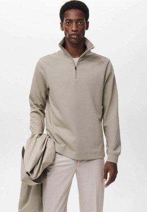 Sudadera beige de media cremallera con tejido texturizado, presenta mangas largas y un corte relajado, combinada con pantalones claros y una chaqueta.
