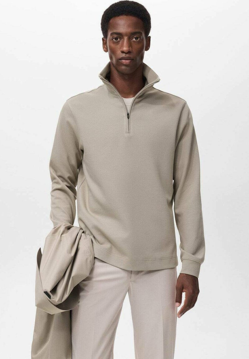 Sudadera beige de media cremallera con tejido texturizado, presenta mangas largas y un corte relajado, combinada con pantalones claros y una chaqueta.