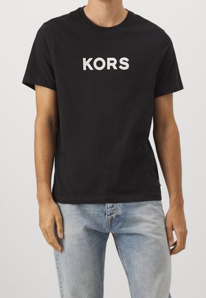 Man wearing a black short-sleeve t-shirt with "KORS" printed in white, paired with light blue faded jeans.