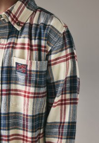 Skjorte i ternet flannel med blå, rød og creme farver. Inkluderer en forlommer med logo-patch og knappet krave. Blød tekstur.