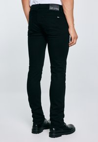 Férfi, aki slim fit fekete farmert és fekete fűzős cipőt visel, kezét nyugodtan oldalt tartva, egyszerű világos háttér előtt áll.